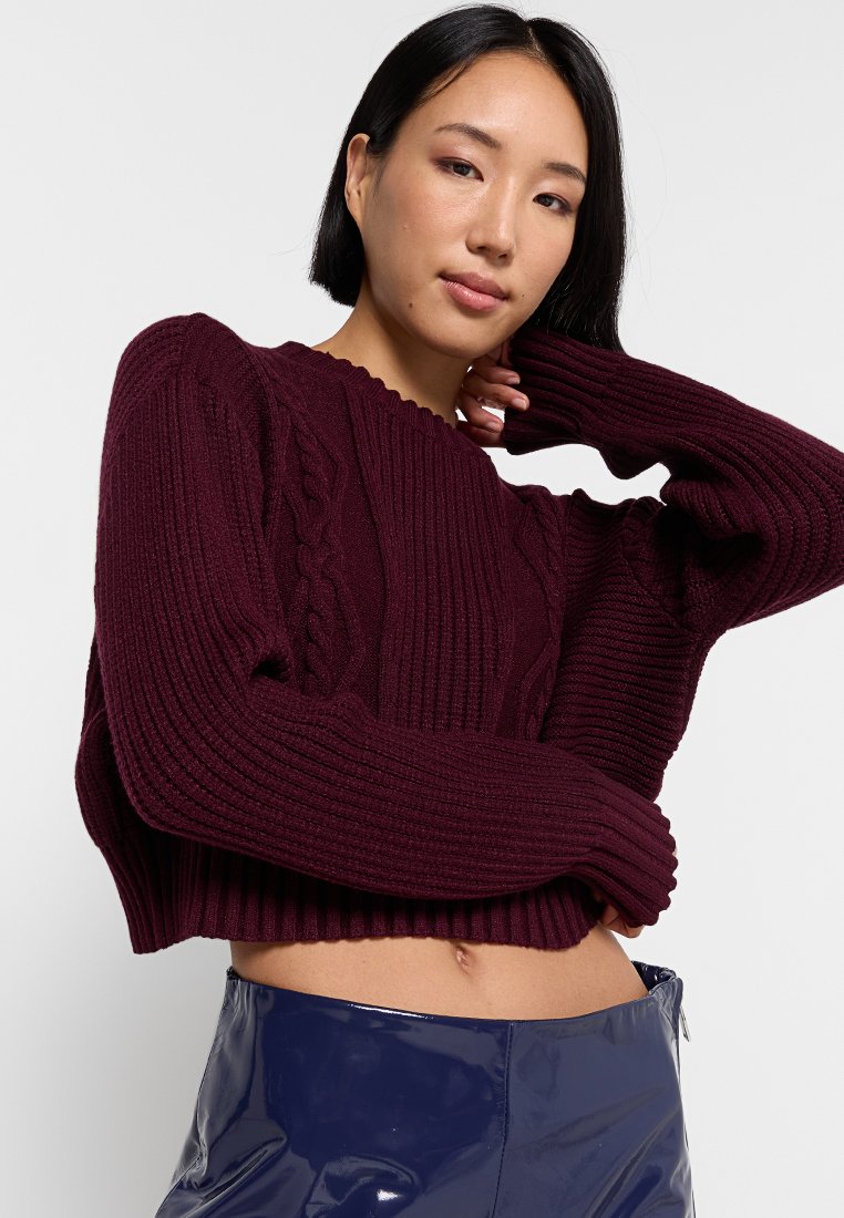Maglione corto a costine color bordeaux con design a treccia texturizzato, maniche ampie e scollatura a onda. Indossato con pantaloni lucidi blu navy.