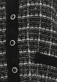Cardigan à carreaux noir et blanc en tissu texturé, doté de boutons noirs brillants avec des accents argentés et d'une poche avant.