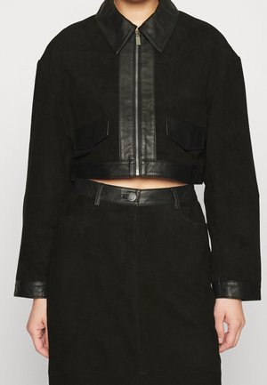 Femme portant une veste courte noire avec bordure en cuir, fermeture éclair devant, poches à rabat et une jupe taille haute assortie avec fermeture par boutons.