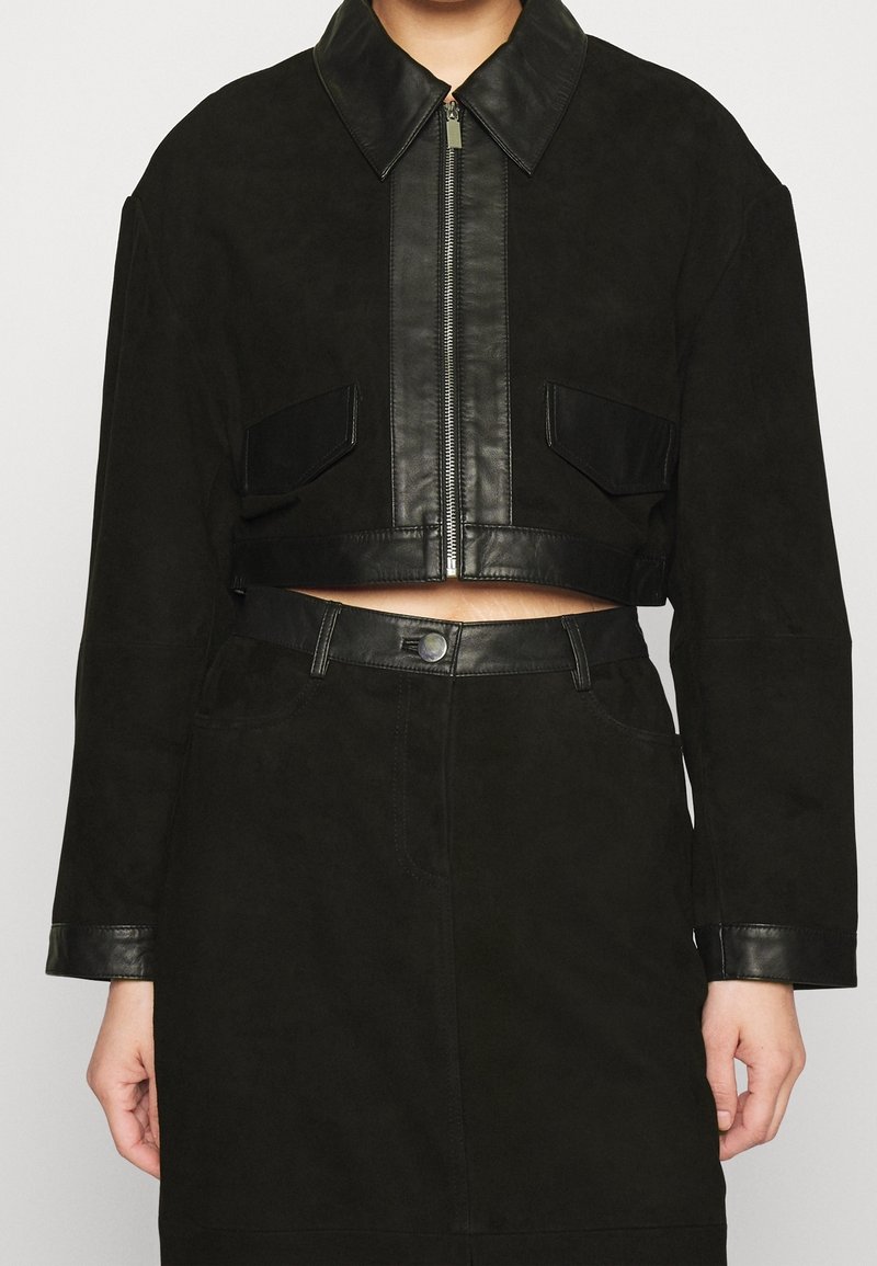 Femme portant une veste courte noire avec bordure en cuir, fermeture éclair devant, poches à rabat et une jupe taille haute assortie avec fermeture par boutons.