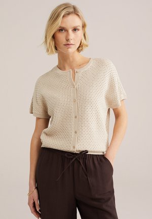 Szőke nő bézs színű rövid ujjú kötött kardigánban és laza, sötétbarna, húzózsinóros nadrágban, zsebébe tett kézzel áll semleges háttér előtt.