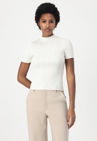 White ribbed short-sleeve top with a high, ruffled neckline. Paired with beige trousers, showcasing a fitted design. Smooth texture.