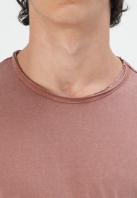 Kurzarmpullover mit Rundhalsausschnitt in einem matten Terrakotta-Farbton, mit gerippter Textur und rohem Rand am Kragen.