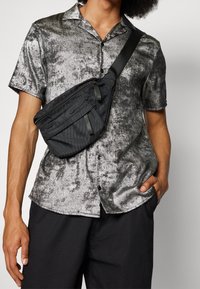 Borsa a tracolla nera in nylon con due tasche con zip. Camicia metallica argento a maniche corte con finitura testurizzata, colletto piatto e bottoni.