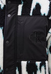 Fekete-fehér polárdzseki türkiz csíkokkal; fekete szövetzsebbel, dombornyomott logóval és patentokkal.