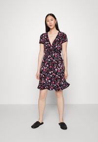 Vestido preto floral com mangas curtas, decote em V e barra rodada. Apresenta flores em tons de rosa e roxo, além de um detalhe na cintura amarrado. Sapatos pretos de slip-on.