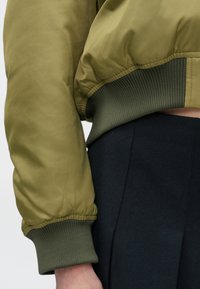 Olivengrønn bomberjakke med en glatt tekstur, ribbede ermer og et lett cropped design, kombinert med et svart høytlivskjørt.