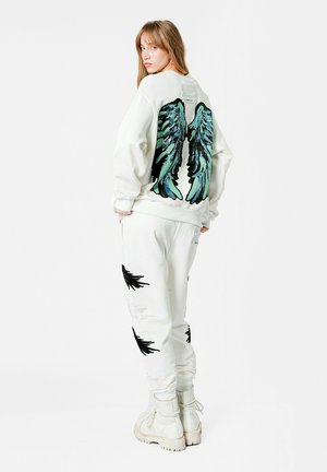 Sudadera blanca con grandes alas multicolores en la parte posterior, combinada con pantalones de chándal blancos con patrones de plumas negras, completada con botas blancas.
