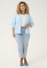 Light blue, knitted cardigan with three-quarter sleeves over a white top, paired with a light denim skirt featuring a front slit and blue sandals.