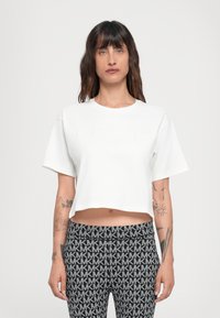 Cropped white t-shirt with short sleeves, featuring embossed "Michael Kors" logo on the front. Worn with patterned black leggings.