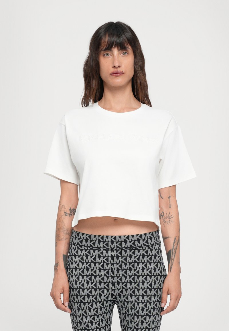 Cropped white t-shirt with short sleeves, featuring embossed "Michael Kors" logo on the front. Worn with patterned black leggings.