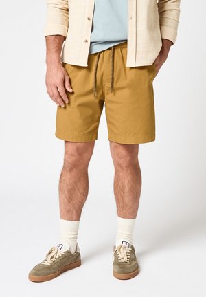 Mann trägt senfgelbe Shorts mit Kordelzug, hellbeiges Hemd, weiße Socken und olivgrüne Turnschuhe, steht vor weißem Hintergrund.