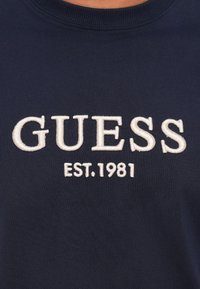 Marineblauwe stof met witte geborduurde tekst "GUESS" boven "EST. 1981" op een kledingclose-up.