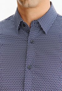 Mężczyzna w granatowej koszuli z białym kwiecistym wzorem i czarnymi guzikami, ukazujący kołnierz i górną część klatki piersiowej.