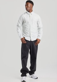 Camisa de botones blanca con finas rayas azules, combinada con pantalones oscuros y holgados. El modelo lleva zapatillas deportivas blancas. Atuendo simple y casual.