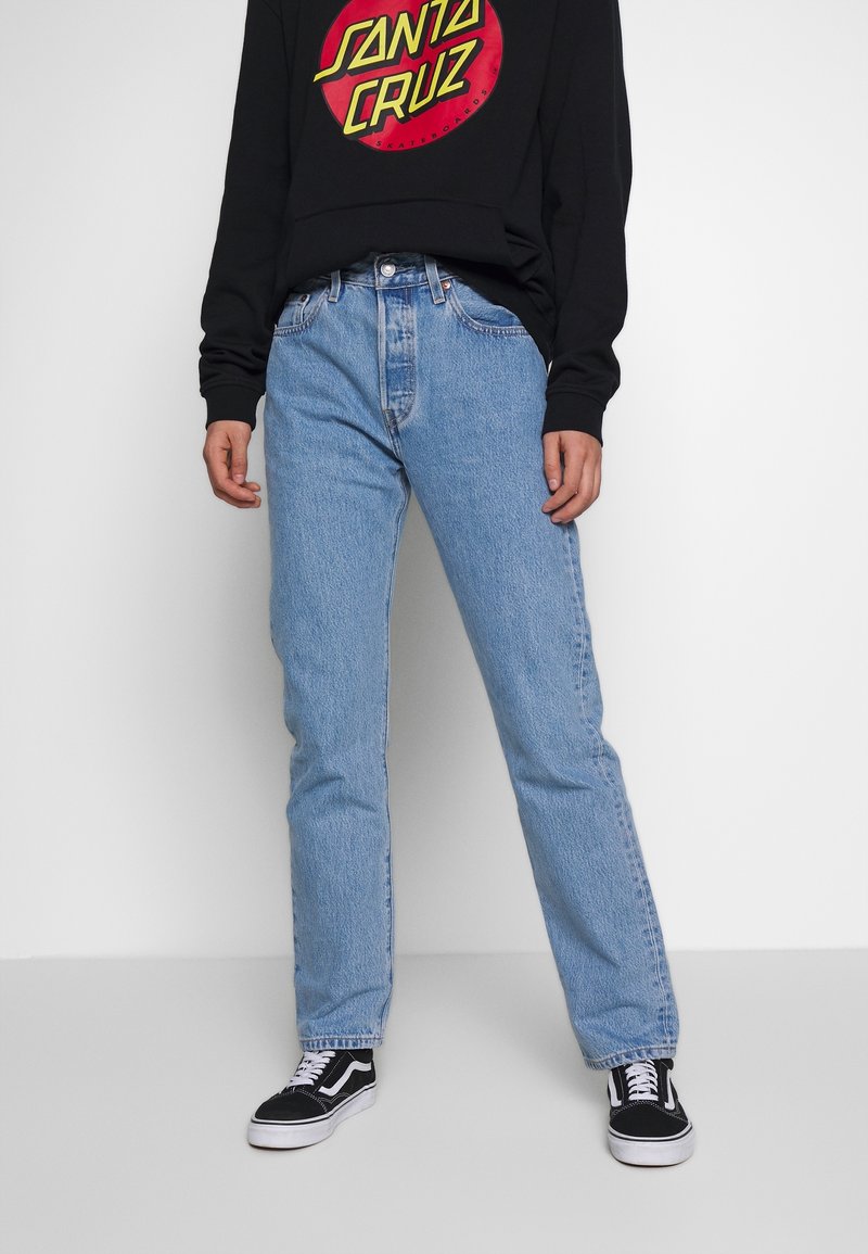 Personne portant un jean droit bleu clair, un sweat-shirt noir Santa Cruz et des baskets à lacets noir et blanc, debout contre un mur blanc.