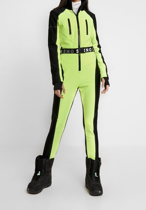 Mujer con un mono de esquí ajustado de color amarillo neón y negro, con botas negras, de pie contra un fondo claro y liso.