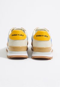 Las zapatillas cuentan con una parte superior de color crema con acentos en azul y marrón, una etiqueta en el talón de color amarillo y una entresuela brillante en beige. Texto "BIMBA Y LOLA" en el talón.