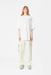 Camiseta blanca de gran tamaño con cuello redondo y mangas cortas, con detalles recortados. Combinada con pantalones color crema y zapatos texturizados.