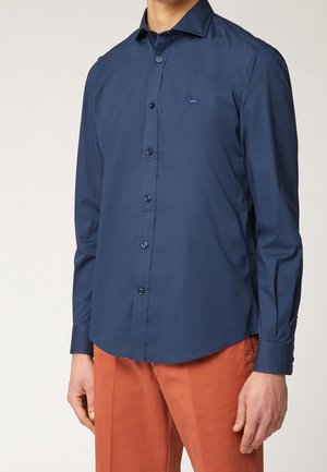 Uomo che indossa una camicia blu navy con bottoni e un piccolo logo ricamato, e pantaloni color ruggine, in piedi su uno sfondo semplice.