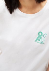 Camiseta blanca con el logo verde "ARMEDANGELS" en el pecho, llevada por una persona con pelo oscuro hasta los hombros.