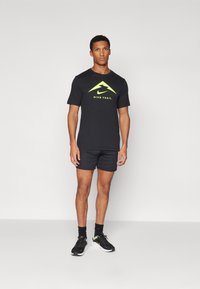 Svart kortermet Nike Trail t-skjorte med gult logo, kombinert med svarte shorts og svarte treningssko. Enkel design, sporty utseende.