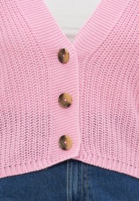 Rozā adīta veste ar V-veida kakla izgriezumu, pieci lieli apaļi brūni pogas, ribota mala un vaļīga auduma tekstūra, kombinēta ar zilo denimu.