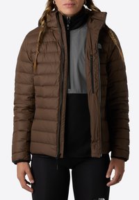 Brązowa kurtka puchowa z zamkiem z przodu, poziomym pikowaniem, przypiętym kapturem i bocznymi kieszeniami, założona na szaro-czarną bluzkę.