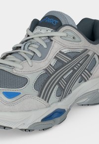 Γκρι και λευκό παπούτσι για τρέξιμο ASICS με μπλε λεπτομέρειες, πλέγμα, μαλακή σόλα και κορδόνια δεμένα σε σταυρωτό μοτίβο.