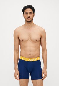 Herrernes Nike boxers i marineblå, med et gult elastisk bælte med Nike-logoet. Glat stof, tætsiddende design og længde til midt på låret.