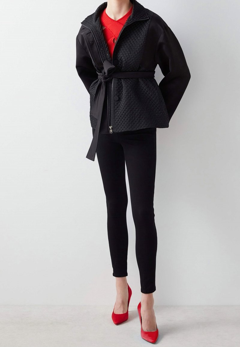 Femme portant une veste matelassée noire avec ceinture, un haut rouge, un pantalon noir ajusté et des talons aiguilles rouges pointus, debout sur fond blanc.