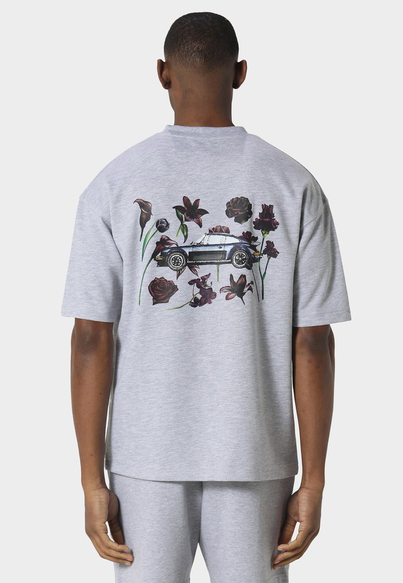 Uomo che indossa una maglietta grigio chiaro con una macchina d'epoca nera circondata da fiori rosso scuro e viola stampata sul retro.