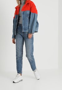 Giubbotto in denim con collo lavorato a maglia rosso, abbinato a jeans azzurri e sneakers bianche. Il giubbotto presenta tasche frontali e orlo sfrangiato.