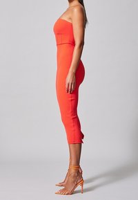 Robe midi rouge sans bretelles, moulante avec un tissu texturé, dotée d'un ourlet évasé et associée à des talons hauts orange à lanières.