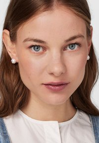 Joven con ojos azules, cabello castaño claro, lleva pendientes de perlas, una blusa blanca y tirantes de mezclilla, mirando directamente a la cámara.