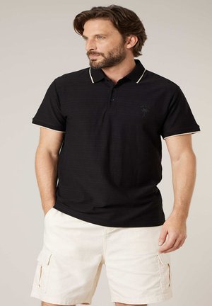 Man met baard, gekleed in een zwart poloshirt met korte mouwen, witte randafwerking en witte cargoshorts, staand met handen in de zakken.