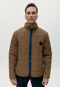 Giacca trapuntata marrone con colletto alto, zip frontale e tasca sul petto con una finitura rotonda nera. Indossata sopra una camicia blu.