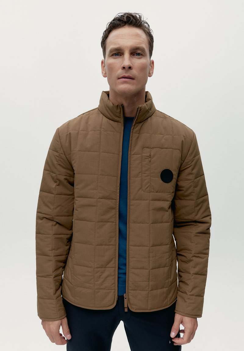 Giacca trapuntata marrone con colletto alto, zip frontale e tasca sul petto con una finitura rotonda nera. Indossata sopra una camicia blu.