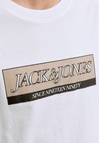 Camiseta de algodón blanca con un gran logotipo rectangular en beige y negro, con el texto "JACK & JONES" y "DESDE MIL NOVECIENTOS NOVENTA."