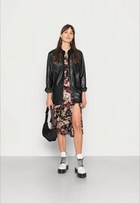 Camisa de couro sintético preta sobre um vestido midi floral com uma fenda na coxa. Botas de plataforma brancas, bolsa preta e brincos de argola completam o visual.