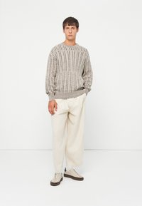 Helles graues Strickpullover mit vertikaler Rippstruktur, lockere Passform. Kombiniert mit cremefarbener, weit geschnittener Hose und beigen Sneakern mit braunen Sohlen.