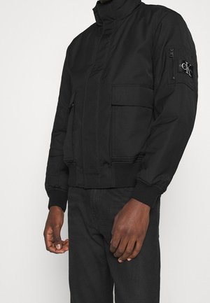 Blouson bomber noir avec fermeture éclair et poches à rabat, poignets et ourlet côtelés, porté par une personne avec un pantalon foncé, patch de marque sur la manche supérieure.