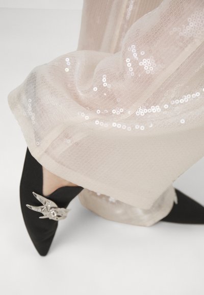 Tacchi neri a punta con un accento d'argento a forma di uccello, abbinati a un tessuto beige leggero, trasparente e decorato con paillettes. Il tessuto si adagia dolcemente attorno alle caviglie.