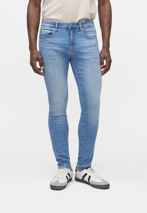 Mand iført lyseblå skinny jeans og hvide sneakers med sorte striber, stående mod en hvid baggrund.