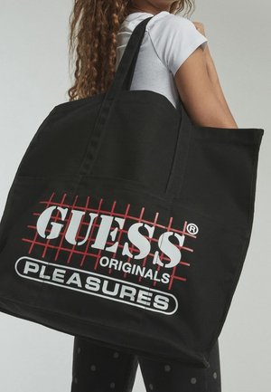 Sac fourre-tout en toile noire avec texte rouge et blanc "GUESS ORIGINALS PLEASURES". Dispose de poignées solides et d'une forme rectangulaire.