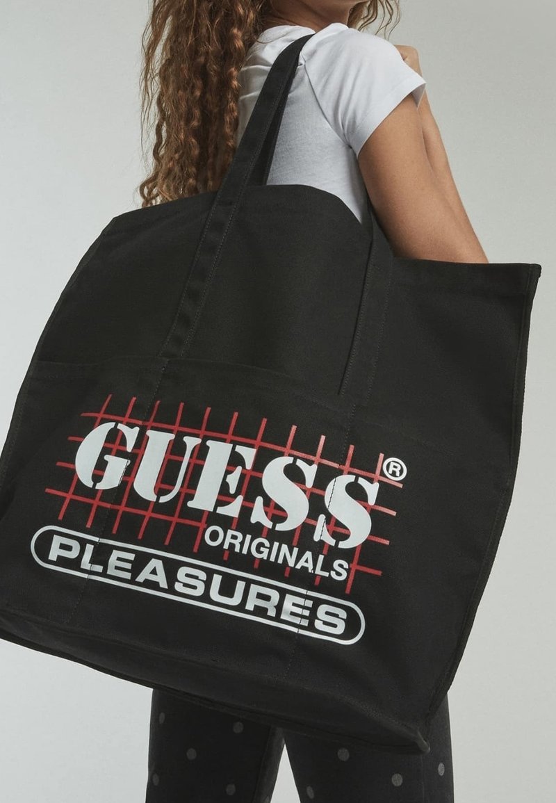 Sac fourre-tout en toile noire avec texte rouge et blanc "GUESS ORIGINALS PLEASURES". Dispose de poignées solides et d'une forme rectangulaire.