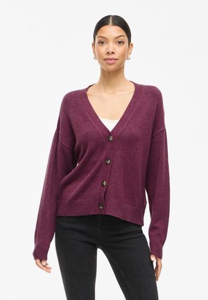 Weinrot cardigan mit V-Ausschnitt, oversized Passform, schwarzen Knöpfen, gerippten Bündchen und Saum. Getragen über einem weißen Oberteil und kombiniert mit schwarzen Jeans.