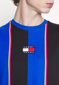 Tricou din bumbac cu dungi verticale îndrăznețe în albastru și negru, accentuat cu linii roz și galbene, și cu un patch logo Tommy Jeans cusut.