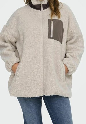 Vrouw draagt een lichtbeige sherpa fleecejack met een ritssluiting aan de voorkant, een donker borstzakje met rits en haar handen in de zijzakken.