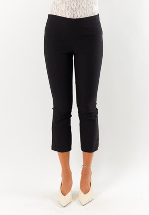 Donna con pantaloni neri corti e tacchi alti bianchi a punta, in piedi su uno sfondo bianco semplice.